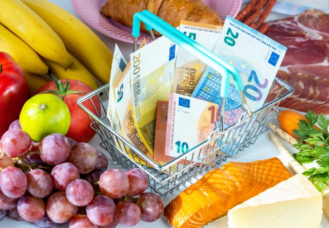 Ein kleiner Korb, in dem Geldscheine liegen, steht zwischen Lebensmitteln wie Obst und Aufschnitt